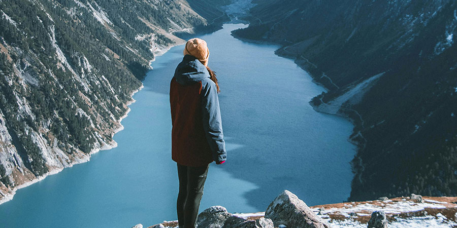 woman wearing winter clothes standing at the edge of a mountain looking at two mountains and a lake in front of her